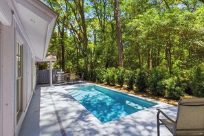 Une piscine avec un espace patio, entourée d’arbres et bordée d’un bâtiment blanc.