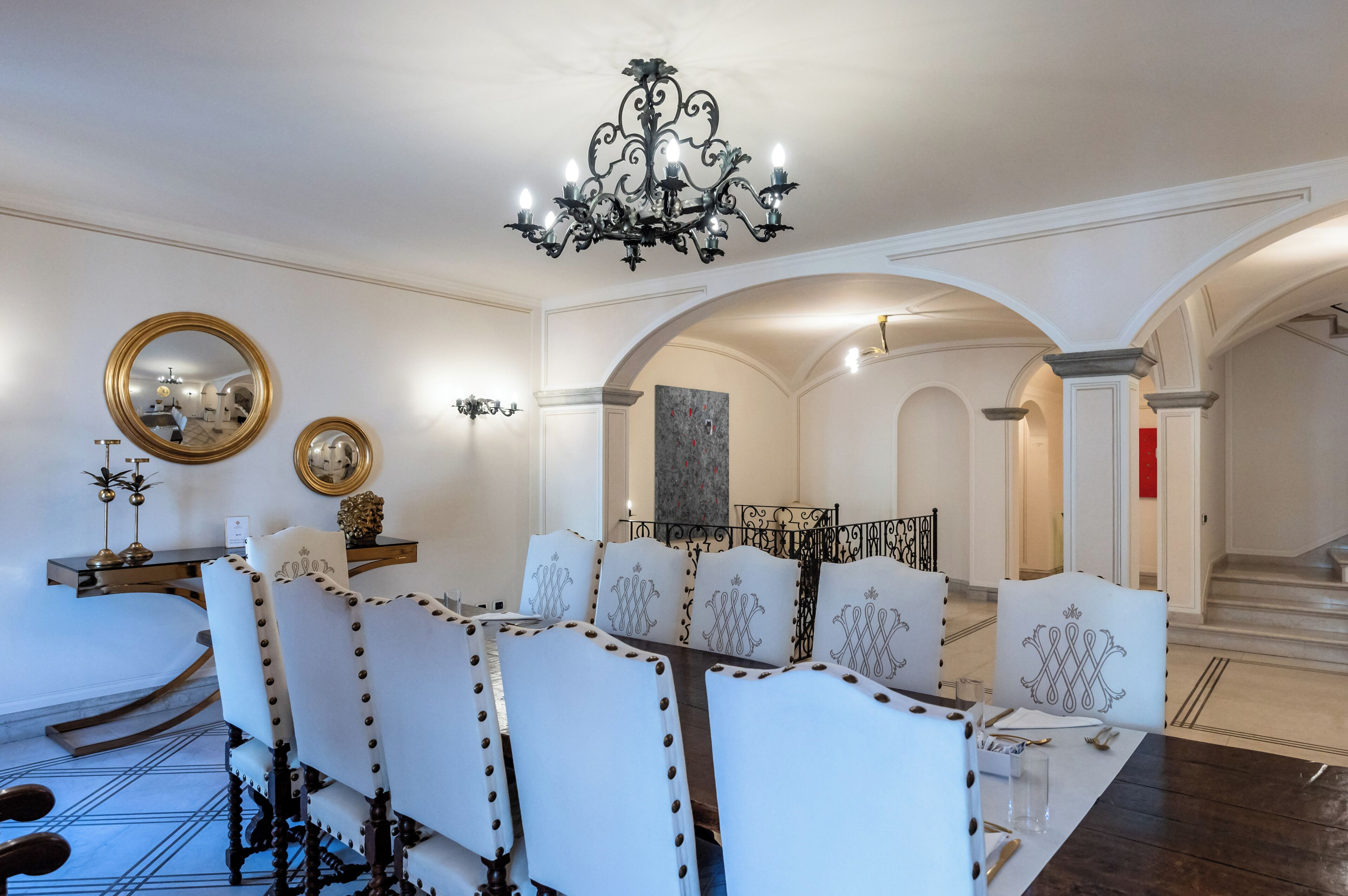 Un coin repas agrémenté d’un lustre, de chaises à revêtement blanc, d’un miroir rond et d’un tableau au mur.