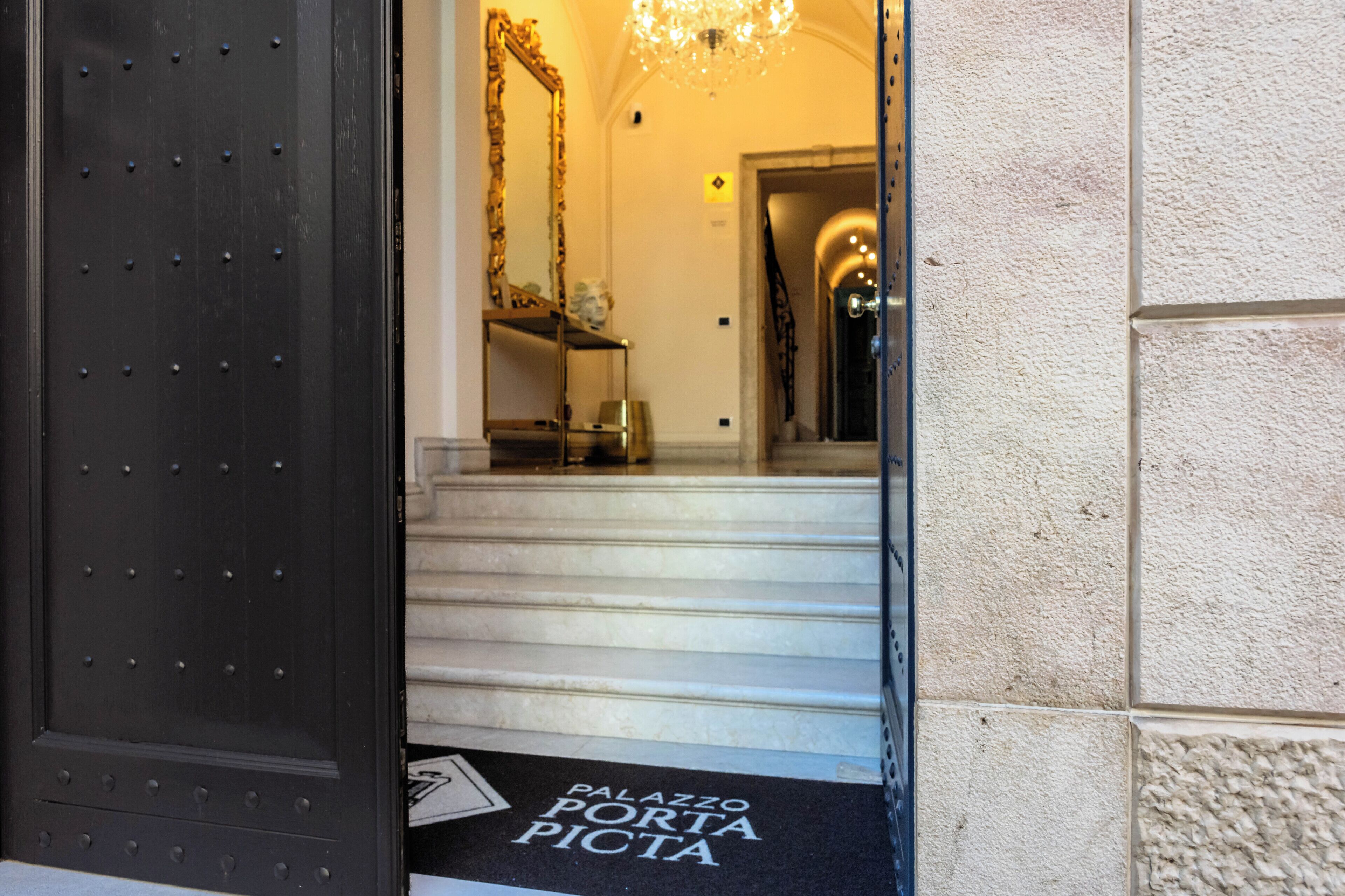 Una scala conduce a un interno maestoso con un lampadario, uno specchio e un cartello con la scritta "Palazzo Porta Picta".