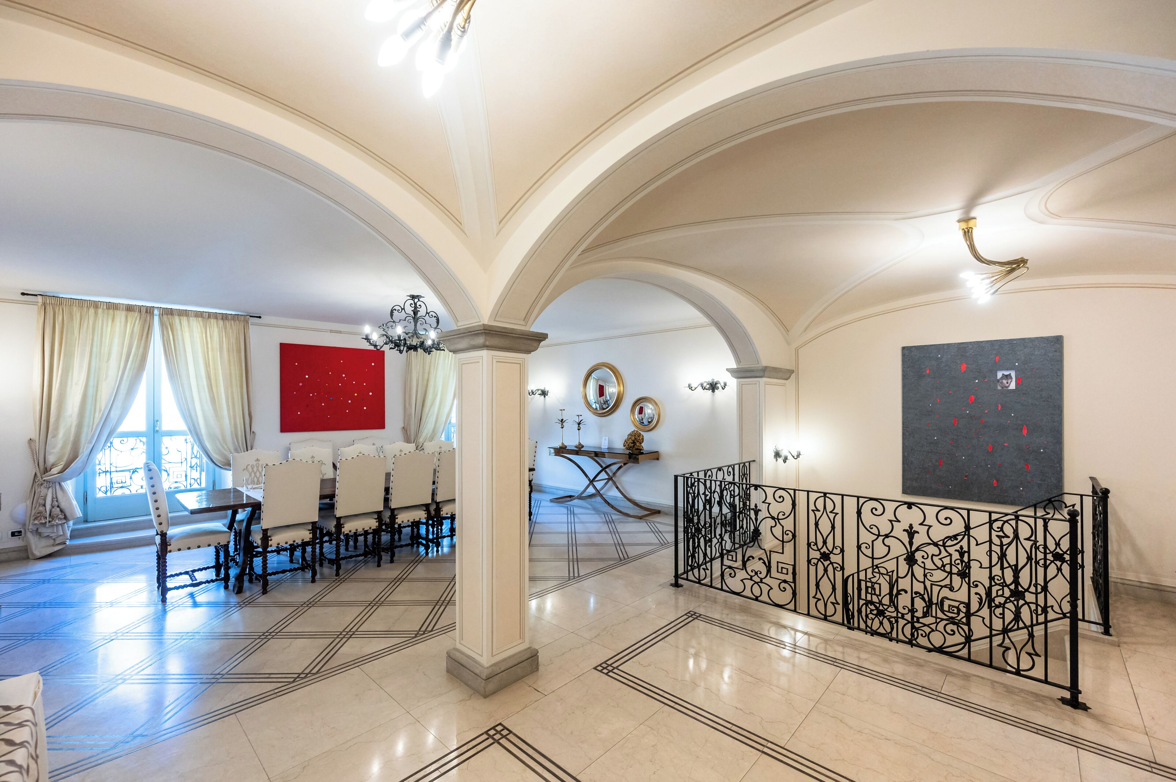 A spacious room with a high ceiling, a dining area with a long table and chairs, and a decorative railing.