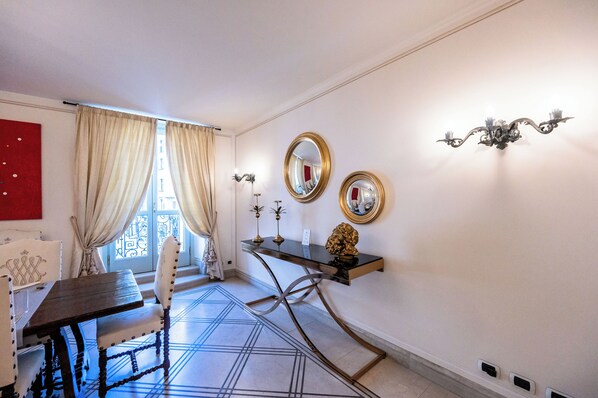 A room with a dining table, chairs, a console table with decorative items, and a view through a window with curtains. - Palazzo Porta Picta (Bergamo)