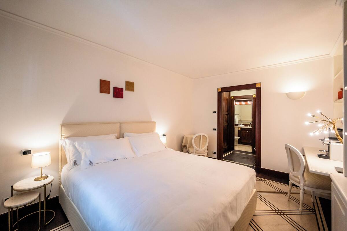 Une chambre d’hôtel avec un grand lit, un bureau avec une chaise, une petite table ronde avec une lampe et un miroir.