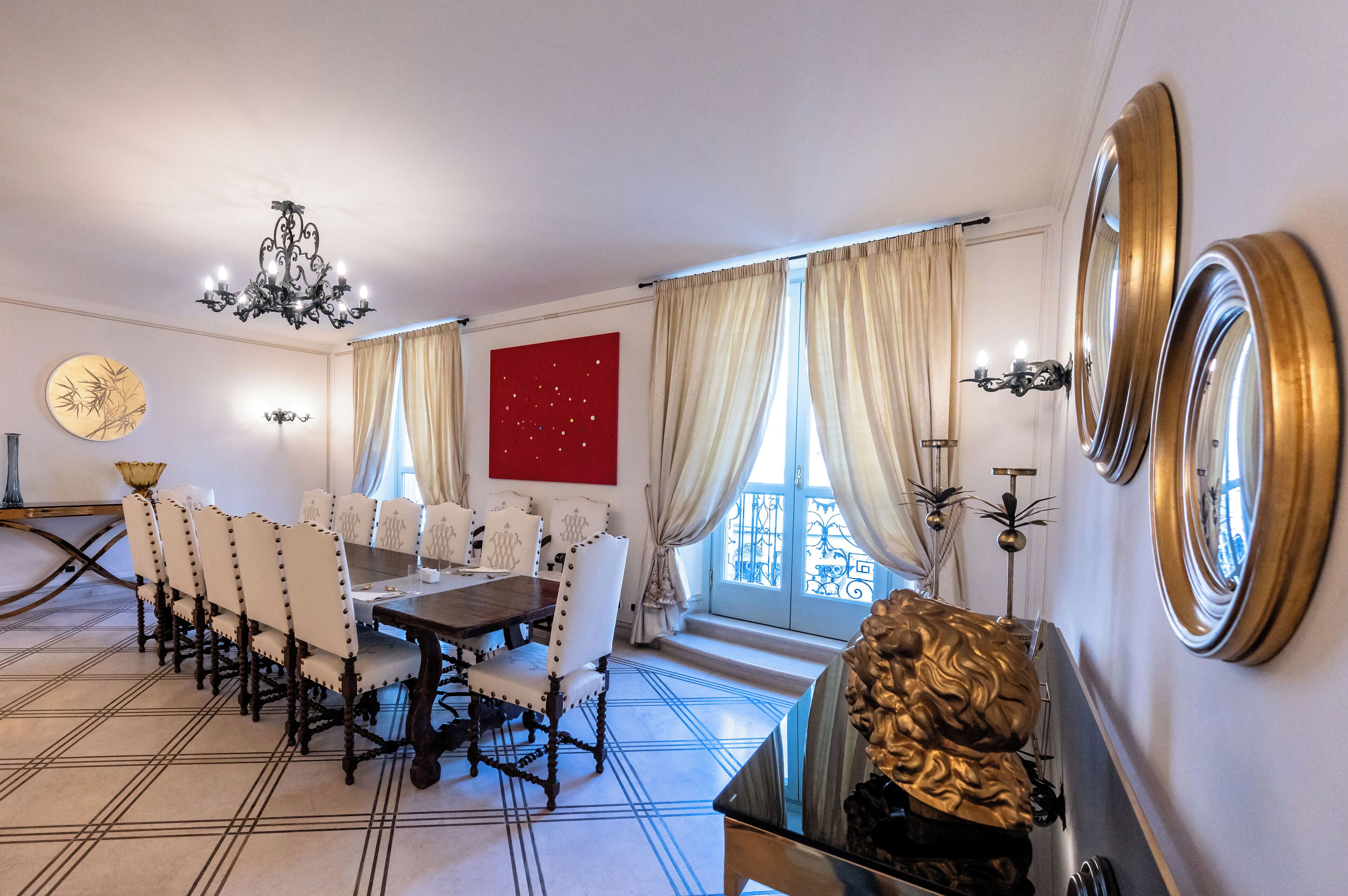A dining room with a long table and chairs, a chandelier, and large windows with curtains.