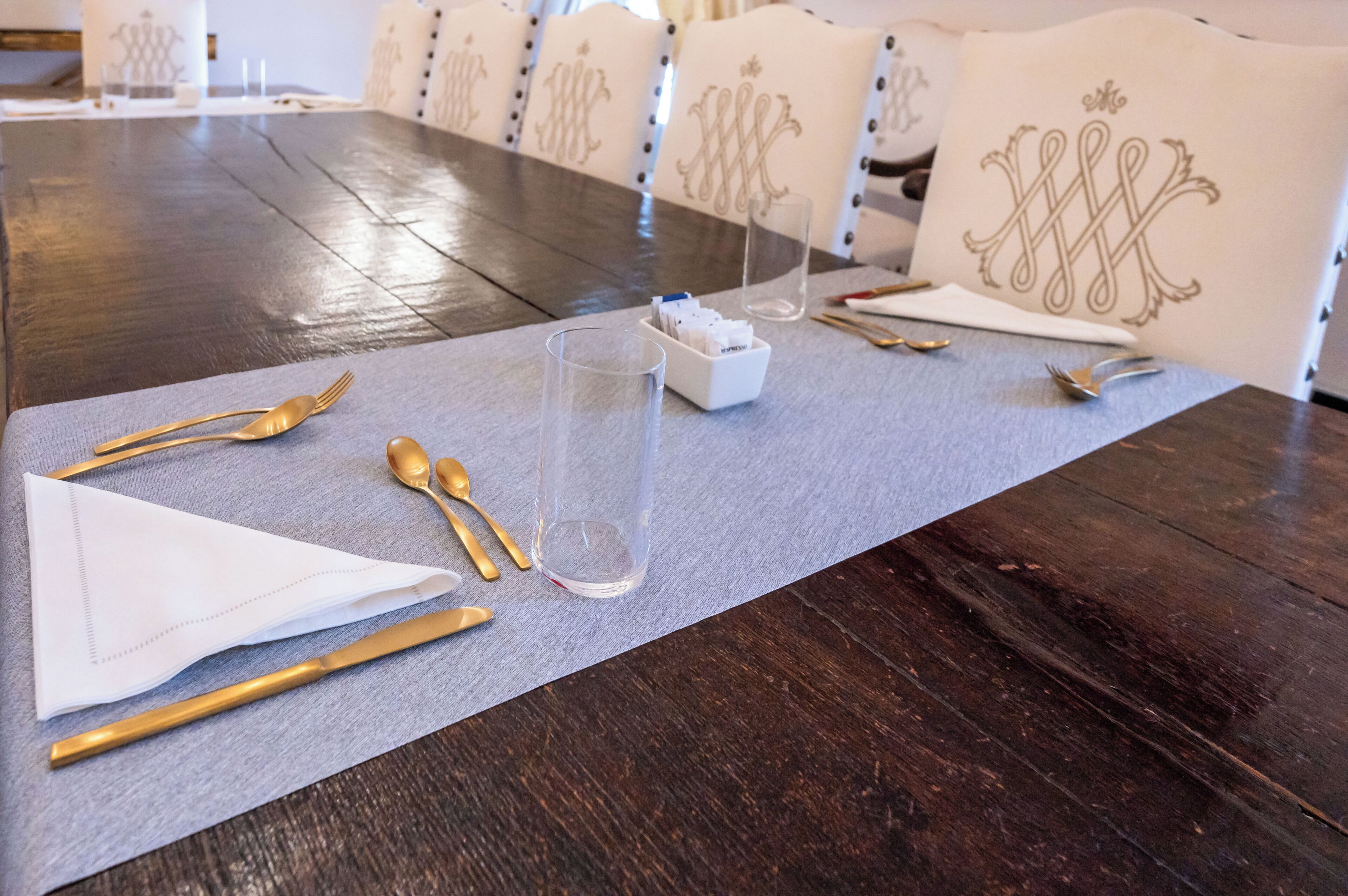 Una mesa de comedor dispuesta con cubiertos dorados, una copa de cristal transparente y una servilleta sobre un mantel individual gris.