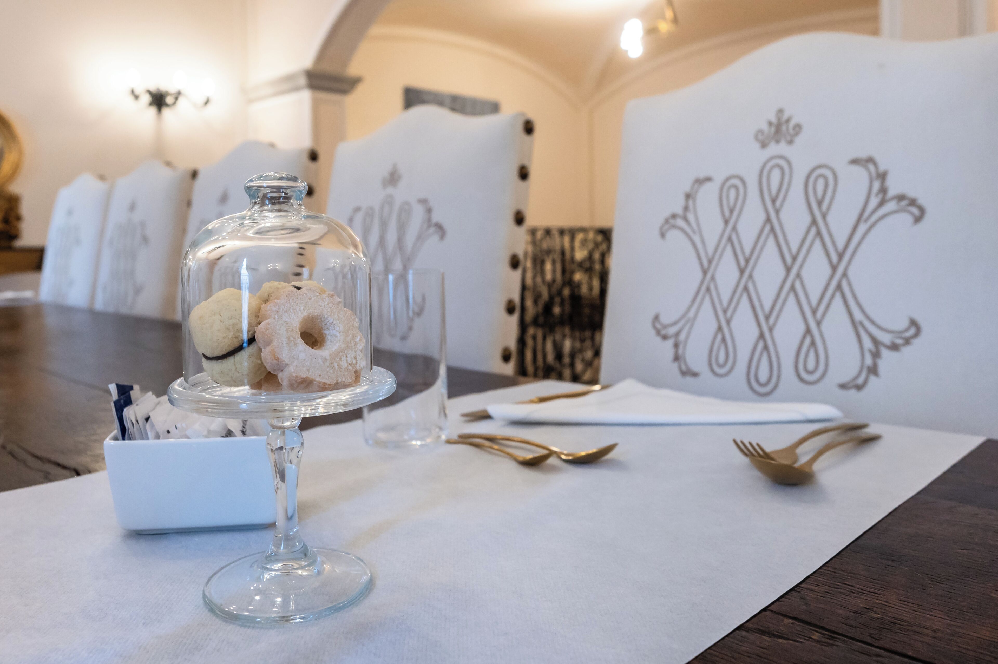 Un tavolo da pranzo con una cupola di vetro che contiene pasticcini, una ciotola bianca piena di dolci e un sottopiatti bianco con un motivo decorativo.