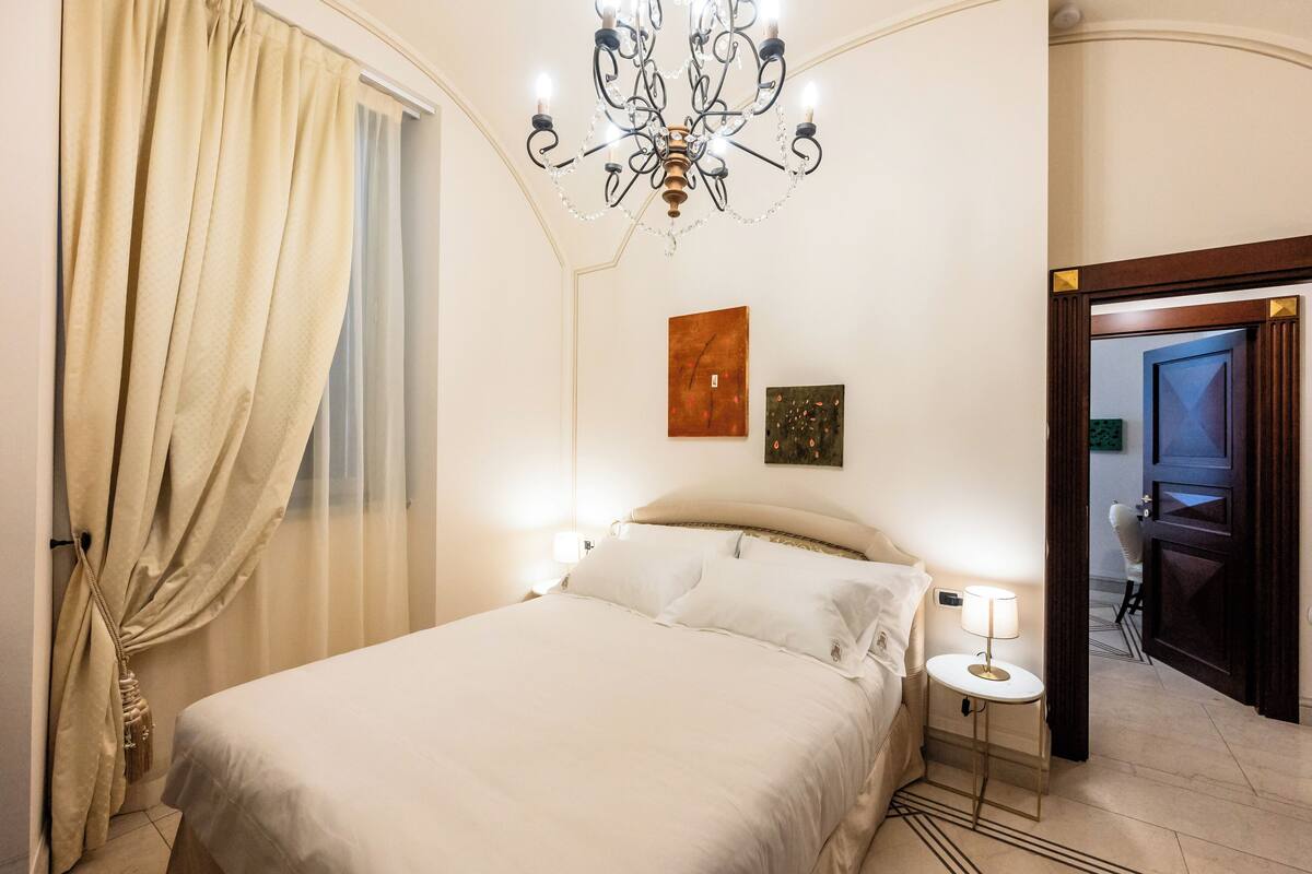 Une chambre d’hôtel avec un lit, un lustre, une table de chevet avec une lampe et des œuvres d’art accrochées au mur.
