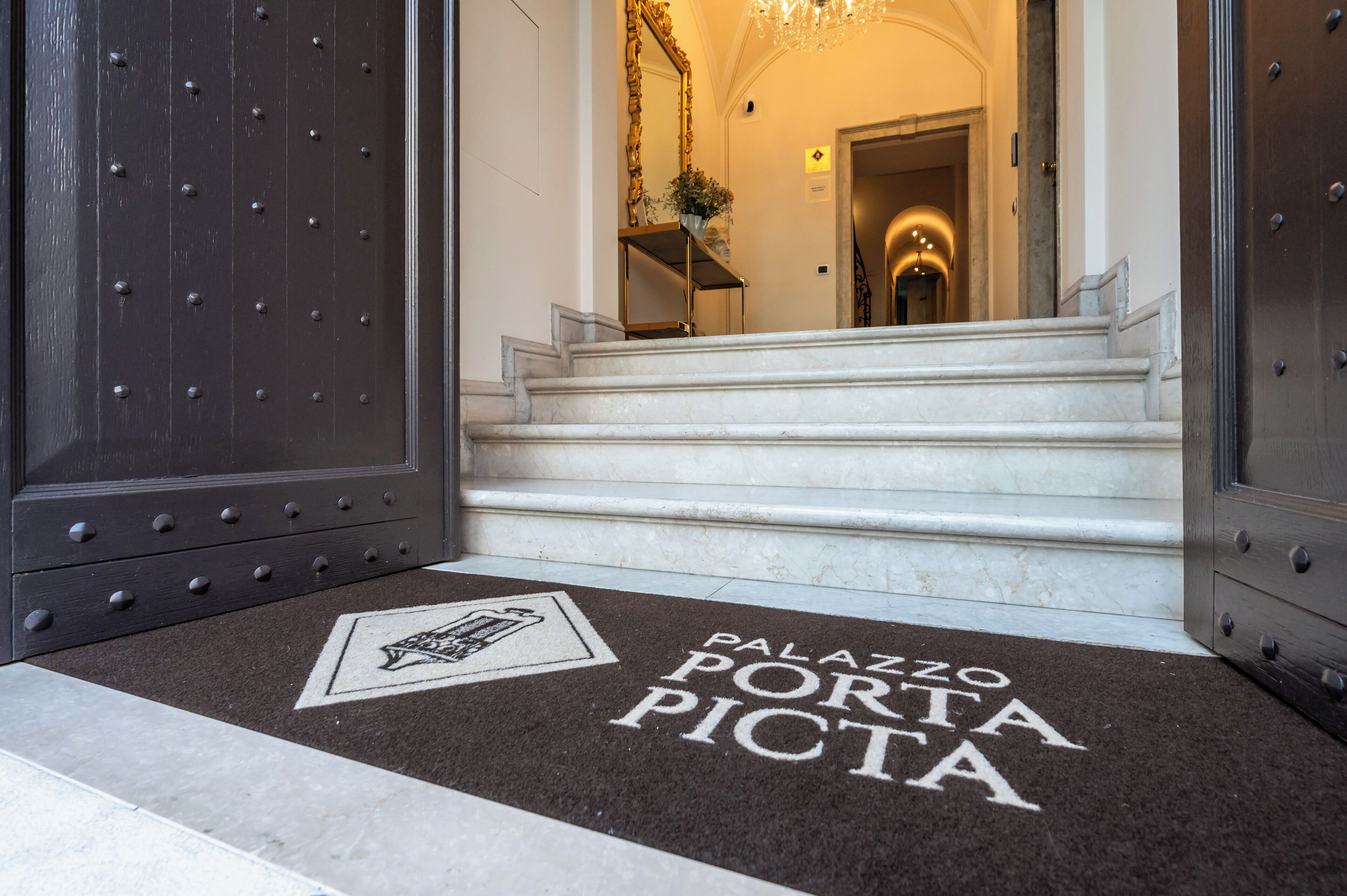 L'entrée d'un hôtel avec un escalier en marbre, un paillasson portant l'inscription « Palazzo Porta Picta » et un grand miroir au mur.