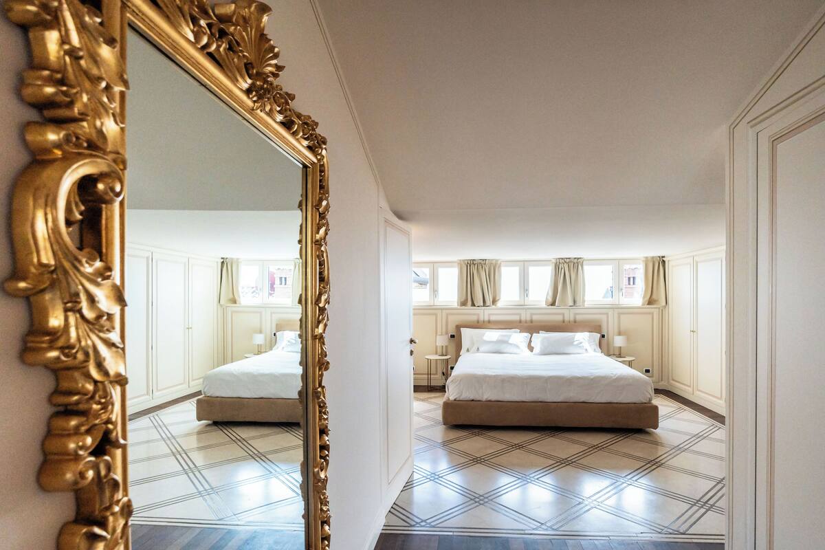 Une chambre d’hôtel avec un grand miroir décoratif, un lit double, une petite table de chevet et une fenêtre avec des rideaux.