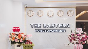 En receptionsdisk med ure, et skilt for The Eastern Hotel & Apartments, og en blomsteropsætning.