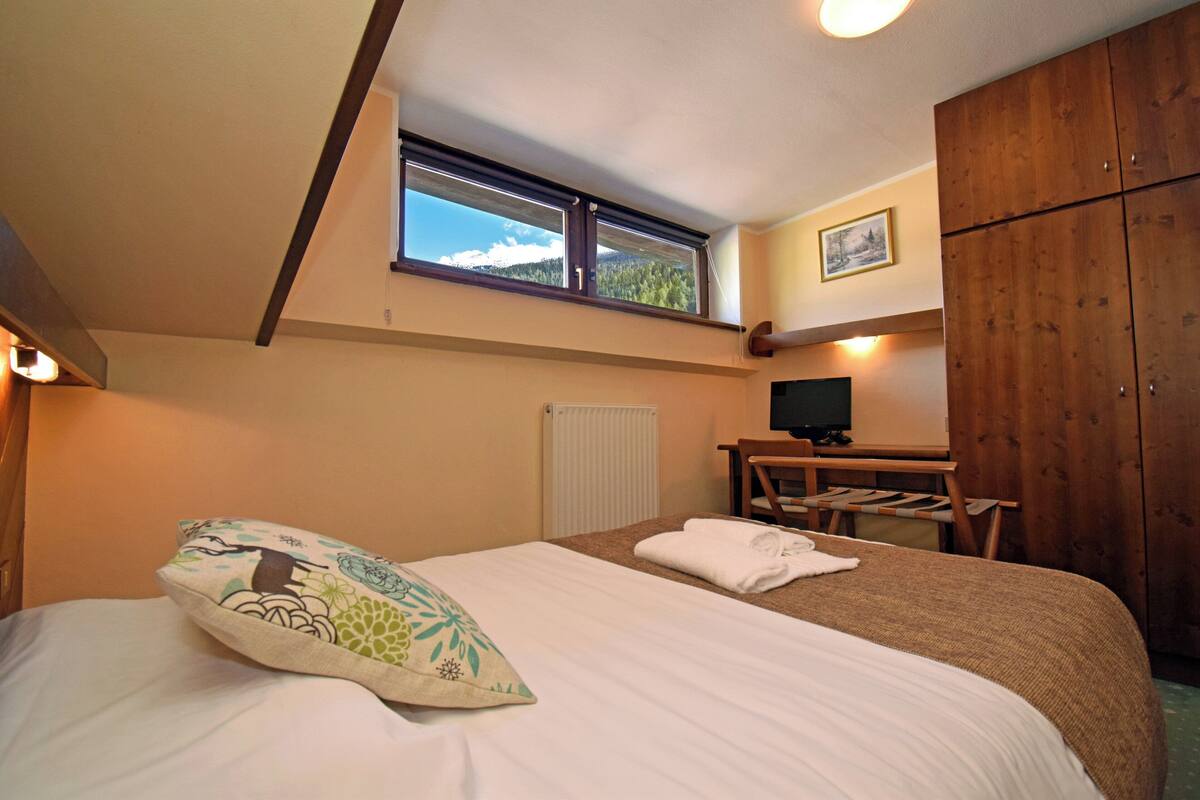 A bedroom with two beds, a wooden wardrobe, a desk with a chair, a television, and a window with a mountain view.