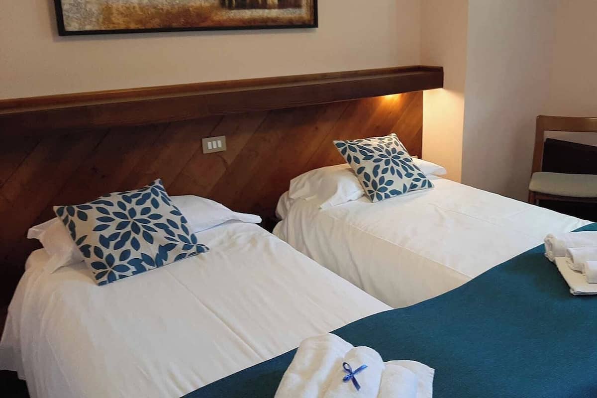 A hotel room with two beds, each with white linens and blue patterned pillows. There is a chair and a folded towel on the bed.