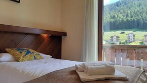A hotel room with a bed, a wooden headboard, a window with a view of a forest and buildings, and a neatly made bed with a folded towel. - Hotel Comelico (Padola)