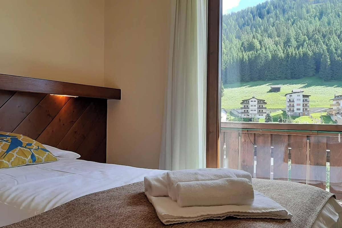 A hotel room with a bed, a wooden headboard, a window with a view of a forest and buildings, and a neatly made bed with a folded towel.