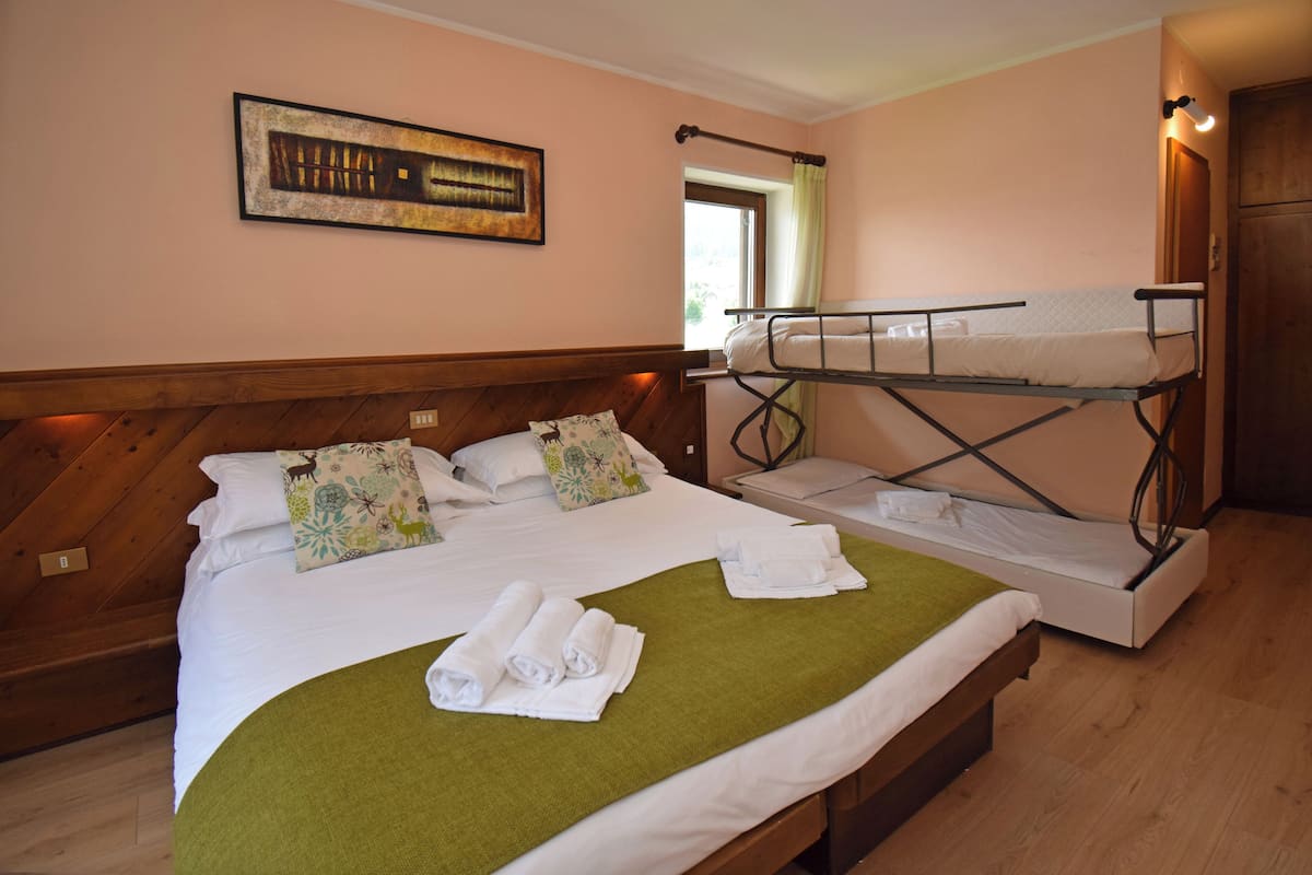A double bed with a green blanket and rolled towels, a bunk bed, wooden headboard, a window with a curtain, and a framed picture on the wall.