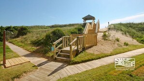 Een houten terras met een trap naar een overdekt gedeelte, omgeven door grasduinen en een heldere lucht.