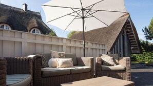 An outdoor seating area with wicker furniture, a large umbrella, and a thatched-roof building in the background. - Thatched roof vacation home Ankerplatz in Fuhlendorf am Bodden (Fuhlendorf)
