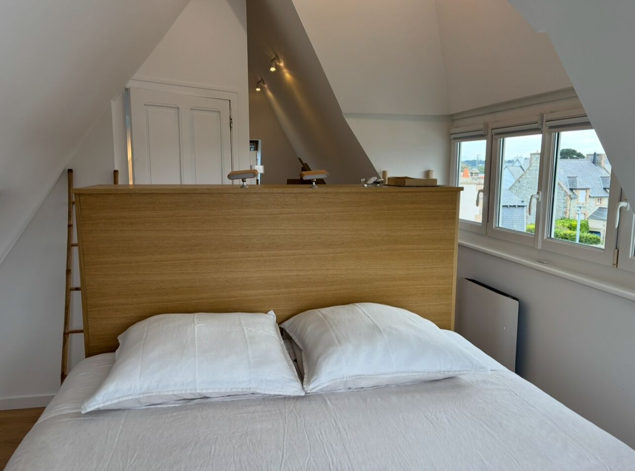 A bedroom with a wooden headboard, white bedding, and a view of the outside through a window.