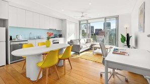 A modern kitchen with white cabinets, a white dining table with yellow chairs, and a city view through large windows.
