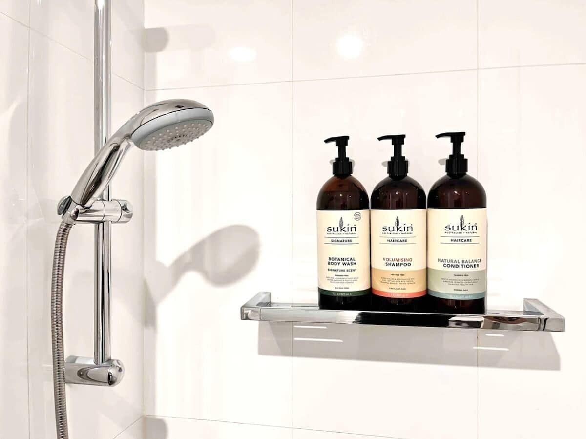 A bathroom with a shower, a glass shelf holding three bottles of Sukin haircare products.