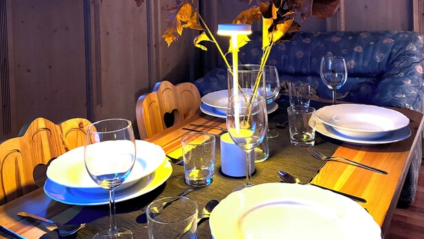 Uma sala de jantar com mesa de madeira posta para refeição, televisão na parede e um sofá com padrão floral ao fundo.