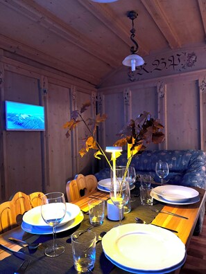 A dining area with a wooden table set for a meal, a television mounted on the wall, and a floral-patterned sofa in the background.