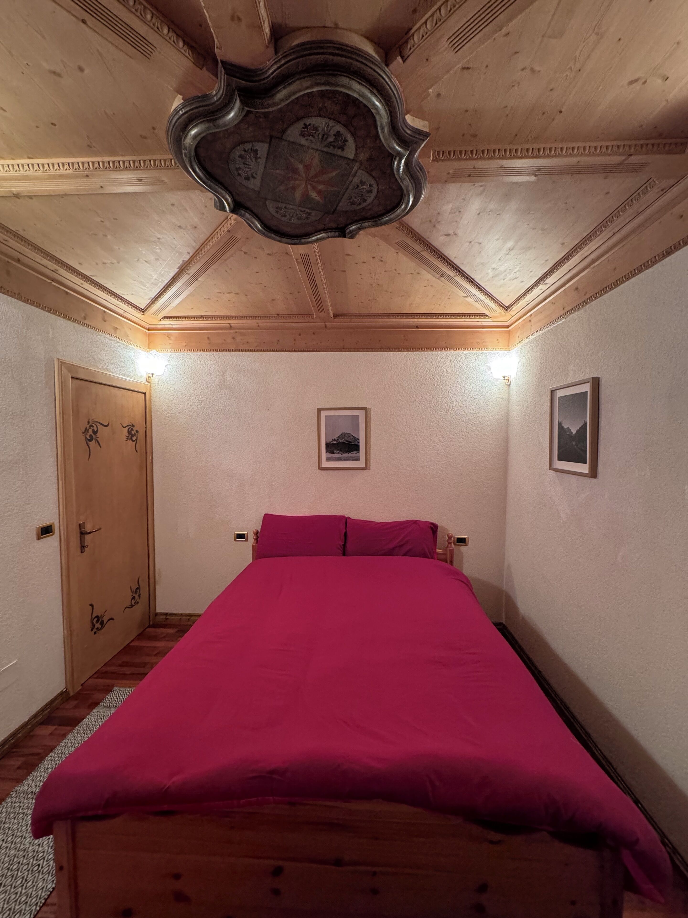 Een slaapkamer met een houten plafond, een bed met een rode sprei, twee ingelijste foto's aan de muur, en een houten deur met kattenmotieven.