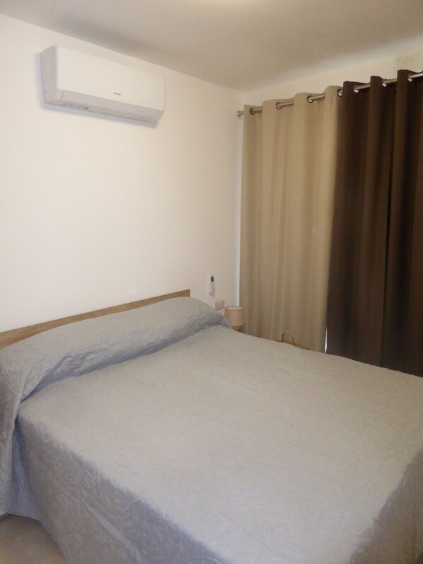 A hotel room with a single bed, a wall-mounted air conditioner, a bedside lamp, and a window with curtains.
