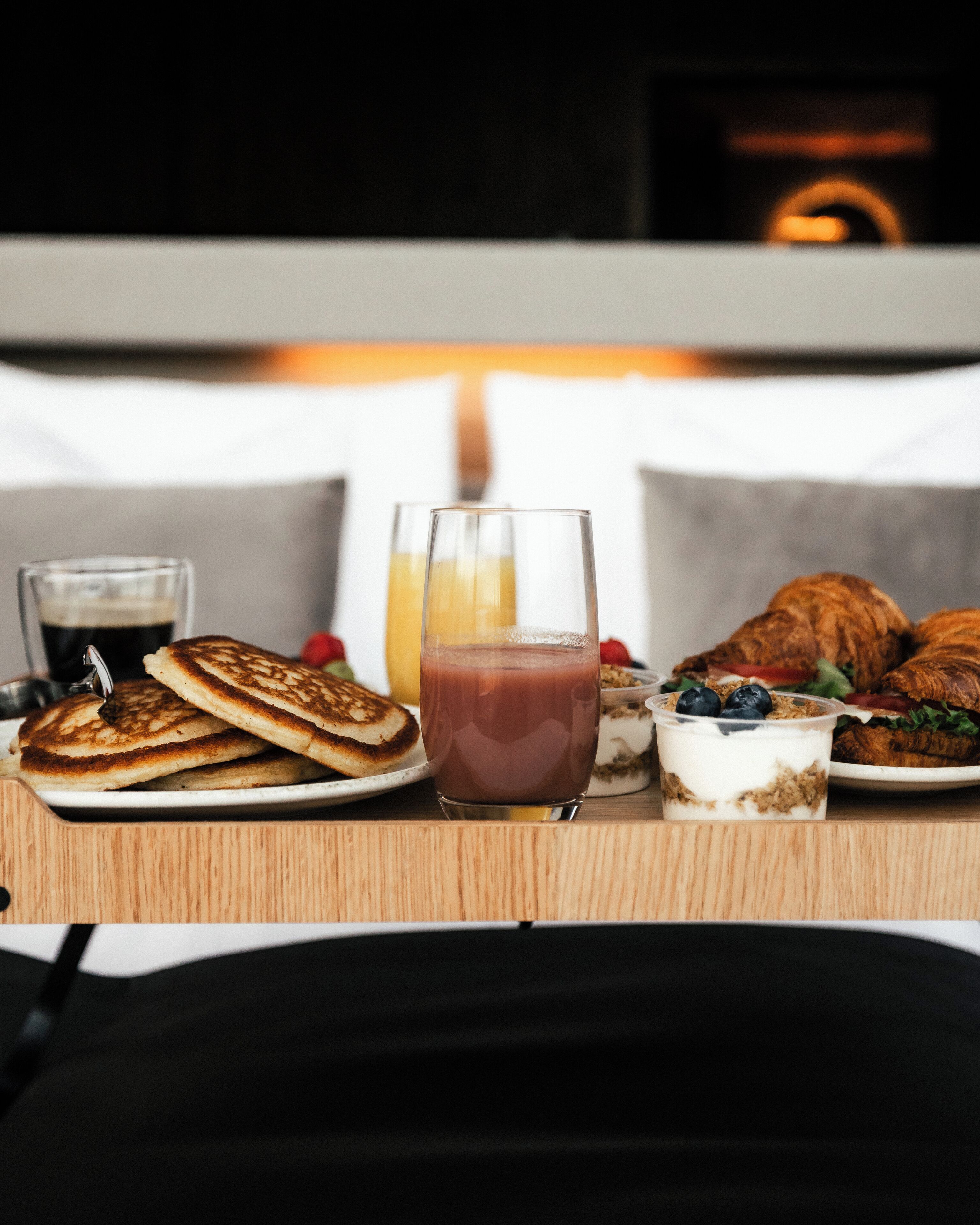A breakfast table with pancakes, a glass of juice, a glass of milk, yogurt with granola, and a croissant.