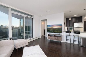 A modern living area with a sofa, a glass sliding door, a painting, and a kitchen with a refrigerator and bar stools.