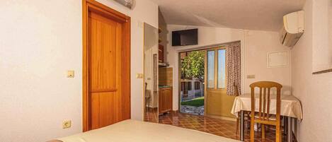 A bedroom with a bed, a wooden door, a small table with a chair, and a view of a garden through an open door.