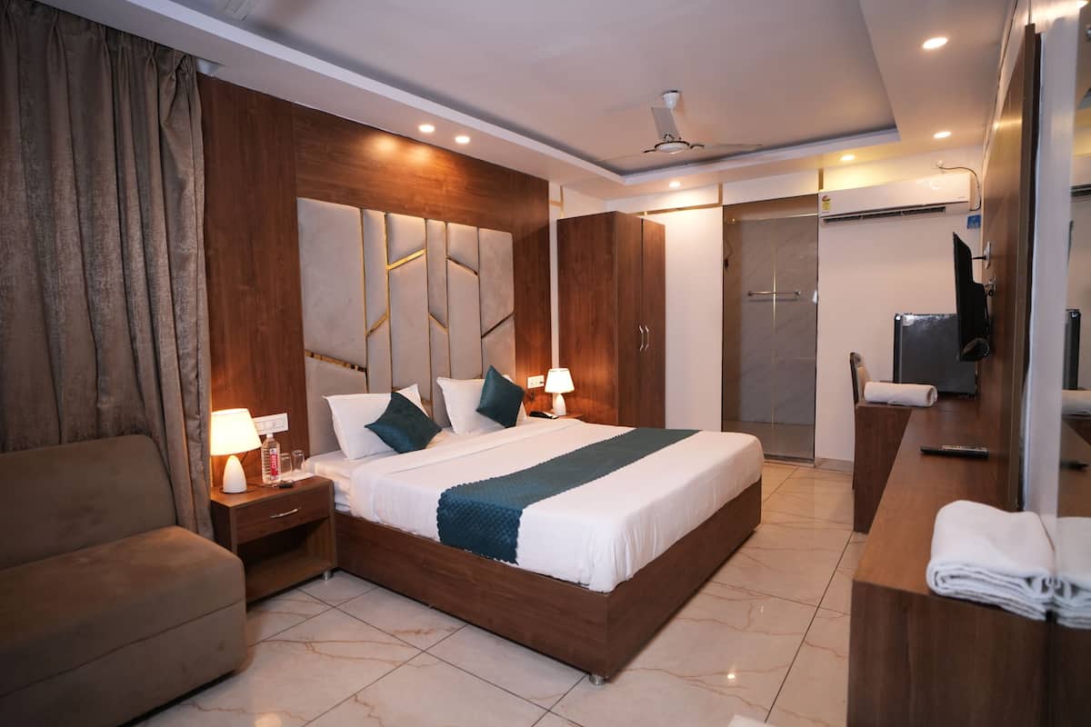 Une chambre d’hôtel moderne avec un grand lit, une tête de lit en bois, des tables de chevet avec des lampes, un canapé, un bureau avec une télévision et un espace douche.