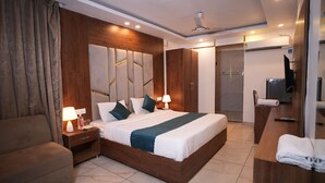Une chambre d’hôtel moderne avec un grand lit, une tête de lit en bois, des tables de chevet avec des lampes, un canapé, un bureau avec une télévision et un espace douche.