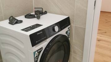 A compact, white Hisense washing machine with a front-loading door, placed in a corner of a room with beige tiled walls and a wooden floor.