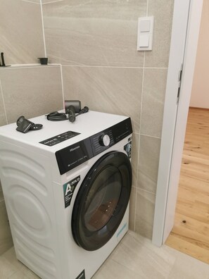 A compact, white Hisense washing machine with a front-loading door, placed in a corner of a room with beige tiled walls and a wooden floor. - Free Parking, Center Location, Nuslesky Pivovar Sunset Balcony Home (Prague)