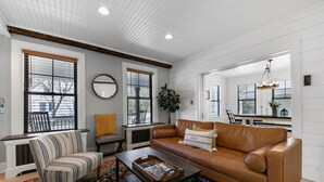 A living room with a brown leather sofa, striped armchair, and a coffee table. There are large windows with blinds, a round mirror, and a potted plant.