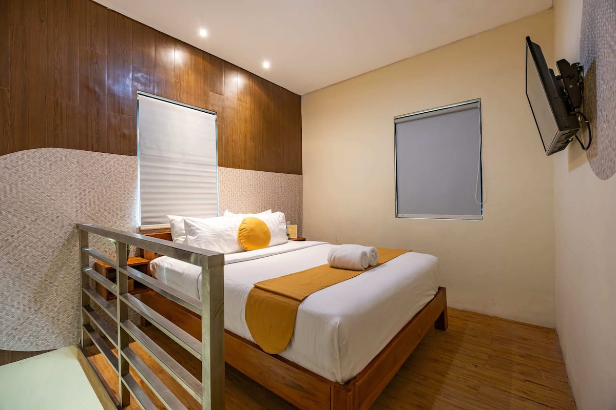 Quarto de hotel com cama de madeira, cabeceira branca, chão de madeira, parede branca e uma janela.