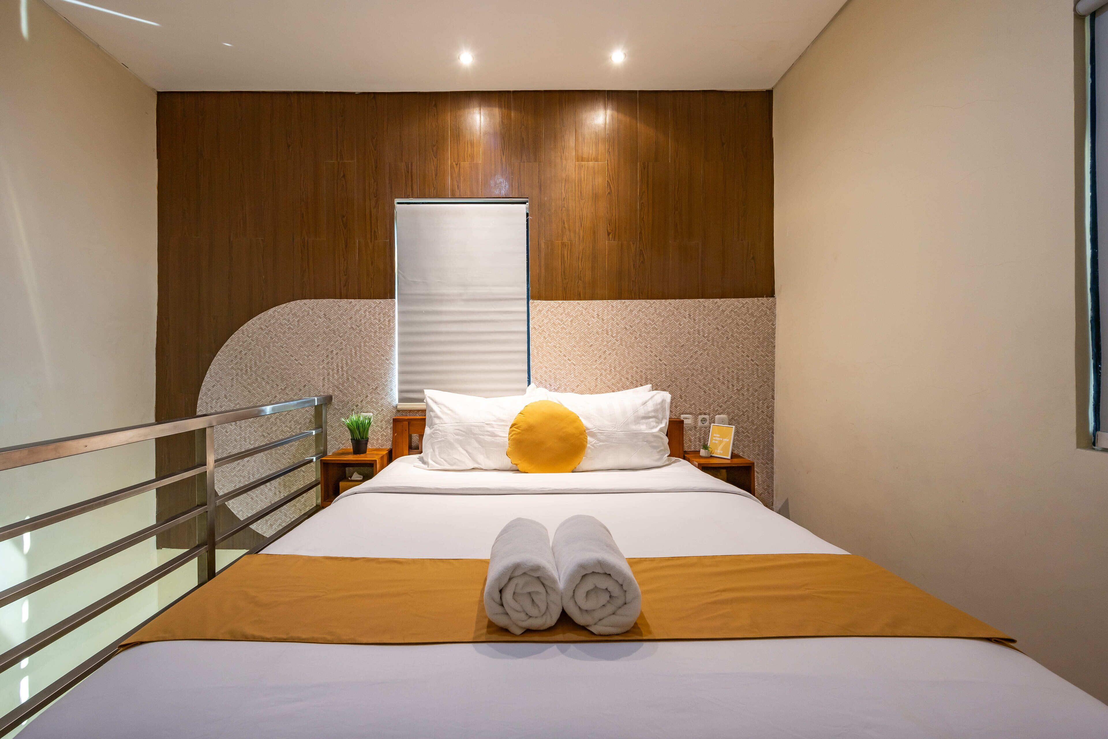 Une chambre d’hôtel avec un lit, une tête de lit en bois, une table de chevet avec une lampe et une fenêtre avec des stores.