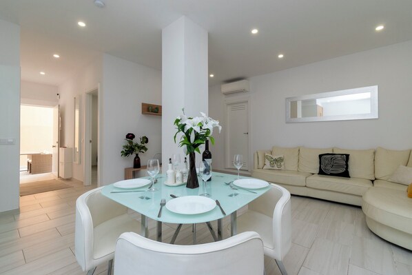 Un comedor moderno con una mesa de vidrio, sillas blancas y un pilar central. Hay un sofá y un espejo en la pared.