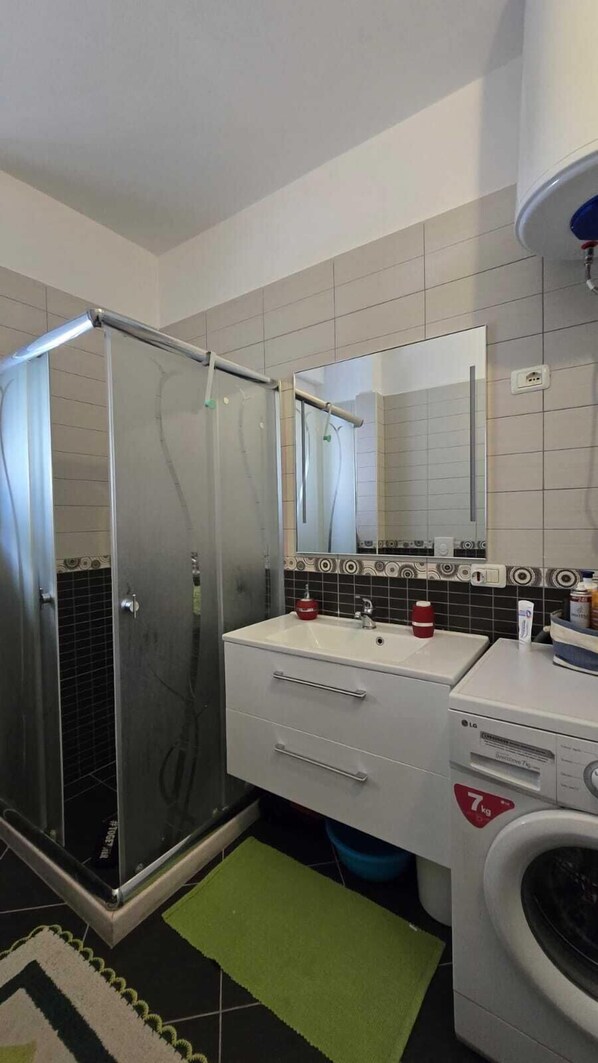 Un baño con ducha de vidrio, lavamanos con espejo, lavadora y una alfombra verde.