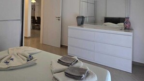 Une chambre à coucher moderne avec un lit, une table de chevet ornée d’un vase, un grand miroir et une commode blanche.
