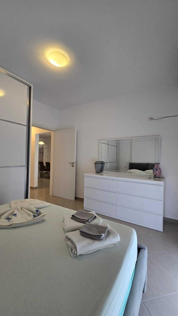 Une chambre à coucher moderne avec un lit, une table de chevet ornée d’un vase, un grand miroir et une commode blanche.