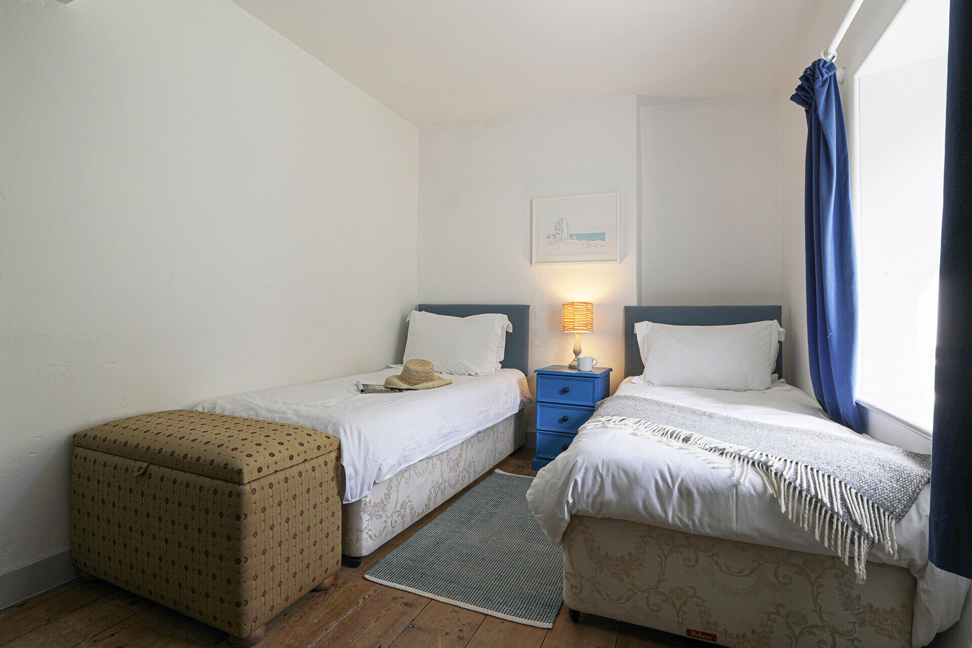 A bedroom with two single beds, a nightstand with a lamp, a small table, a patterned ottoman, and a framed picture on the wall.