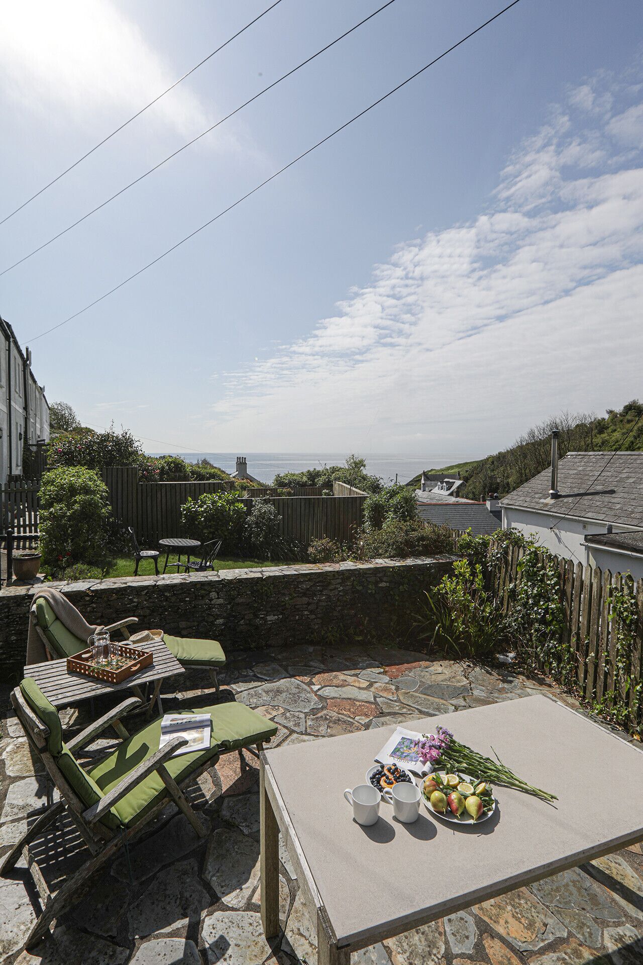 Une terrasse avec un sol en pierre, des chaises rembourrées vertes, une table sur laquelle se trouve une assiette de fruits et un bouquet de fleurs, et une vue sur la mer.