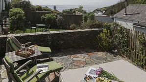 Een terras met stenen vloer, groene kussens op de stoelen, een tafel met een fruitschaal en een bloemenboeket, en uitzicht op zee.