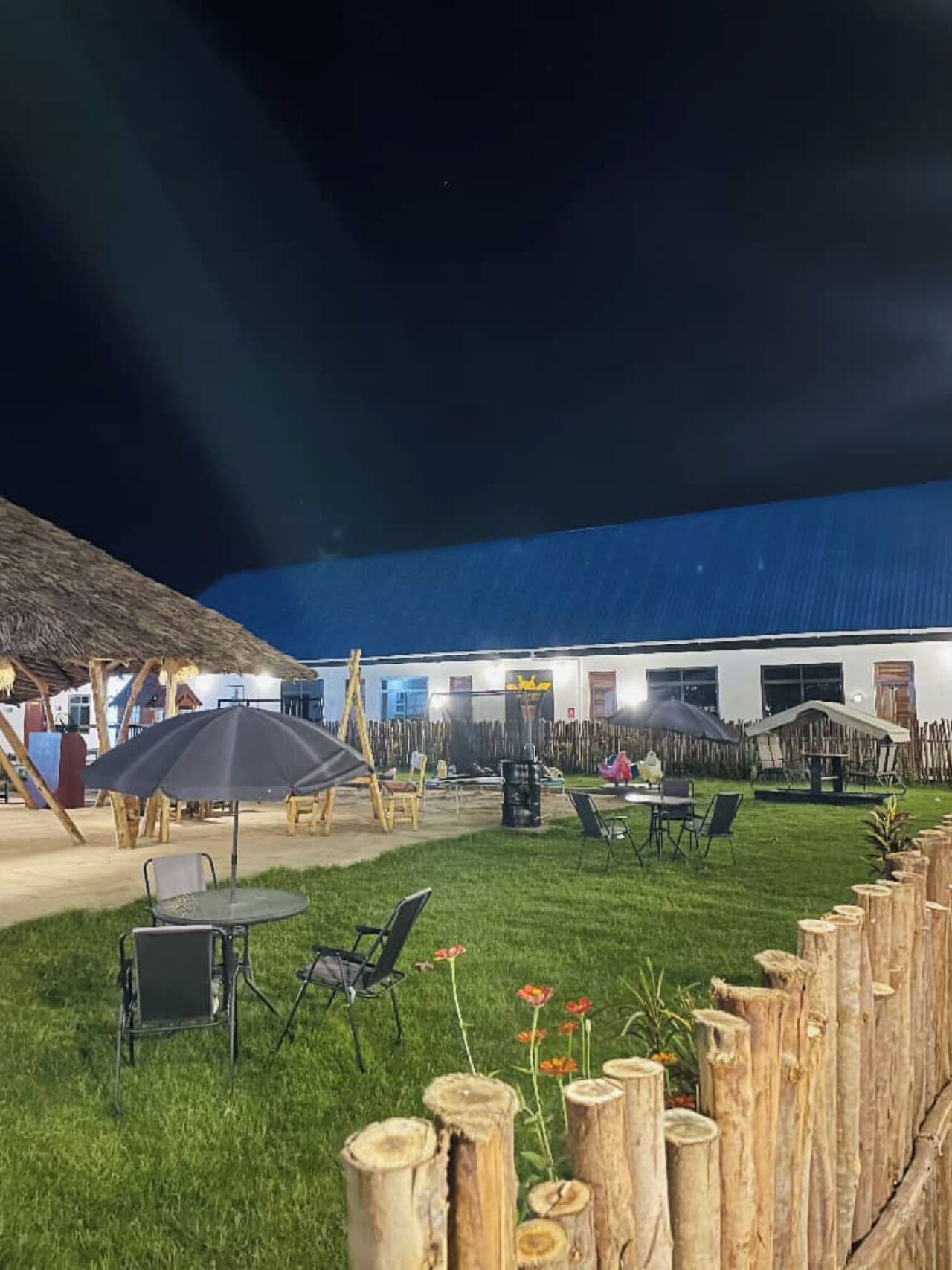 An outdoor seating area with tables and chairs, a thatched-roof structure, and a wooden fence.