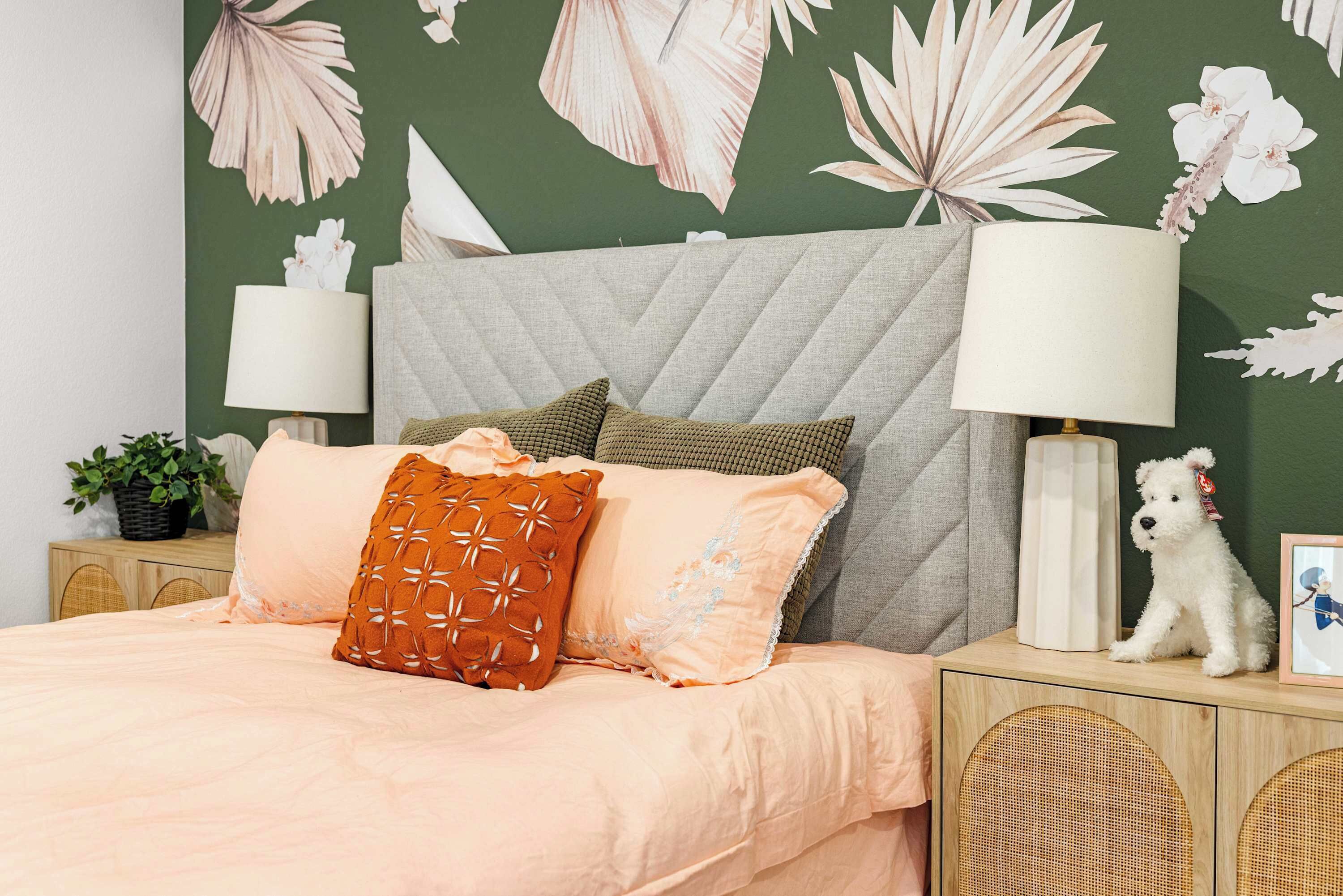 A neatly made bed with a peach-colored bedspread and decorative pillows, a grey upholstered headboard, and a wall with a tropical leaf pattern.