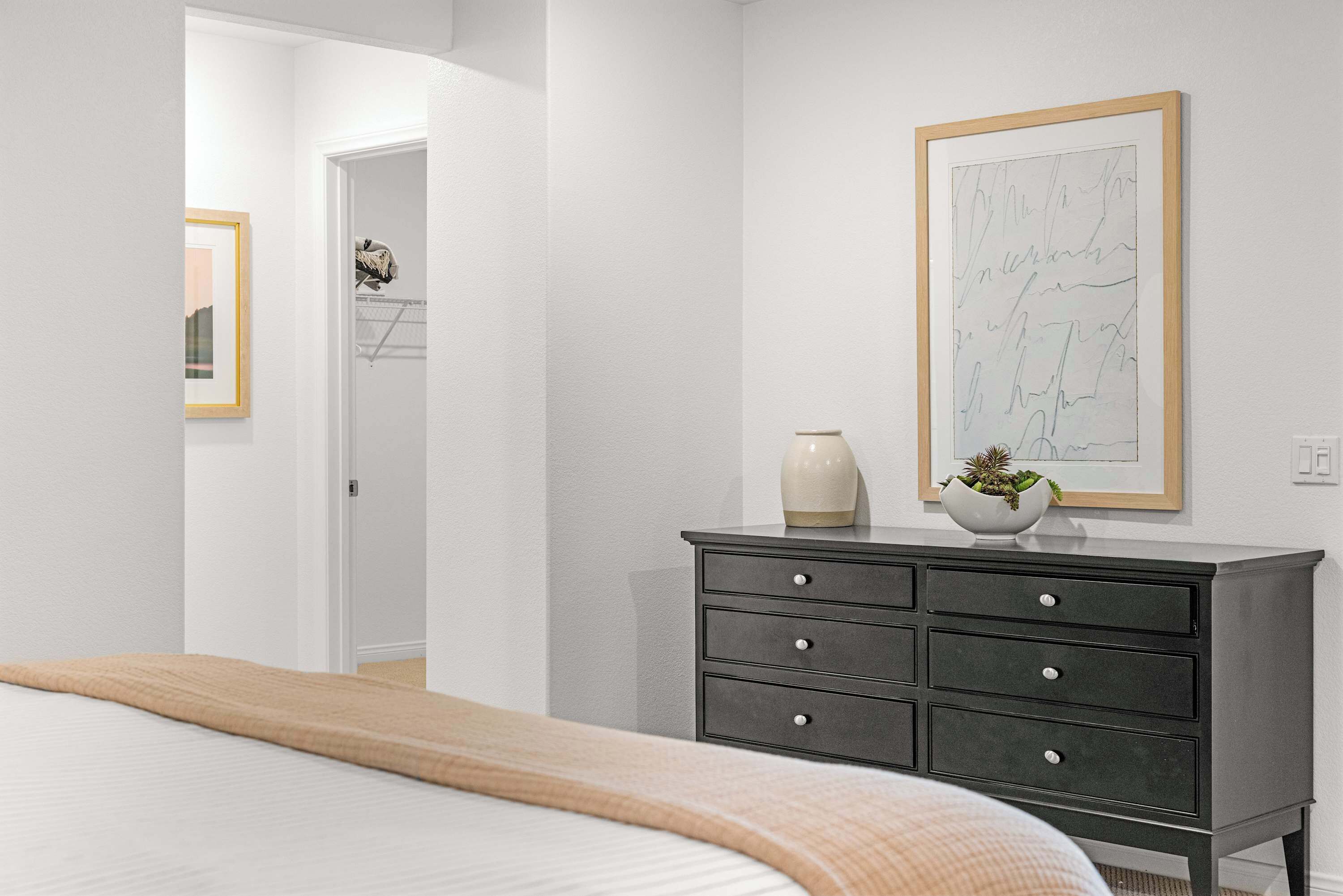 A bedroom with a bed, a dresser, a framed artwork, and a door leading to another room.