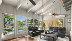 A spacious living room with a large window, a black leather sofa, a coffee table, and a ceiling fan. - @ Marbella Lane - Alpaca Farm House l Pool (Modesto)