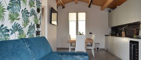 A modern kitchen with a wooden ceiling, a dining area with a blue sofa, and a wall with tropical leaf wallpaper.