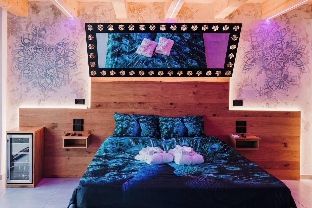 A bedroom with a wooden headboard, a large bed with peacock-themed bedding, two bedside tables with lamps, and a wall-mounted TV.